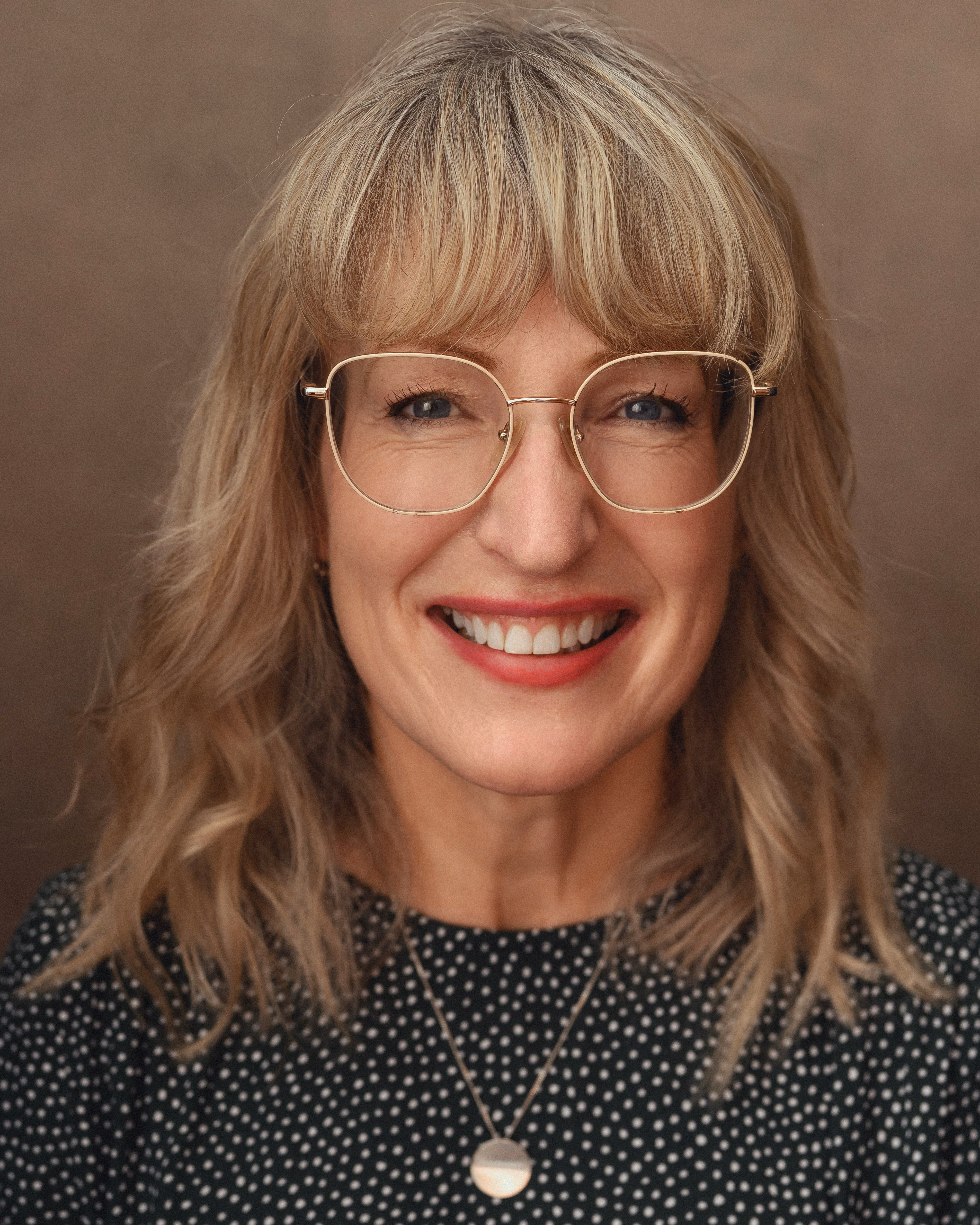 Smiling woman with blonde hair, glasses, red lipstick, wearing a black polka-dot top and round necklace.