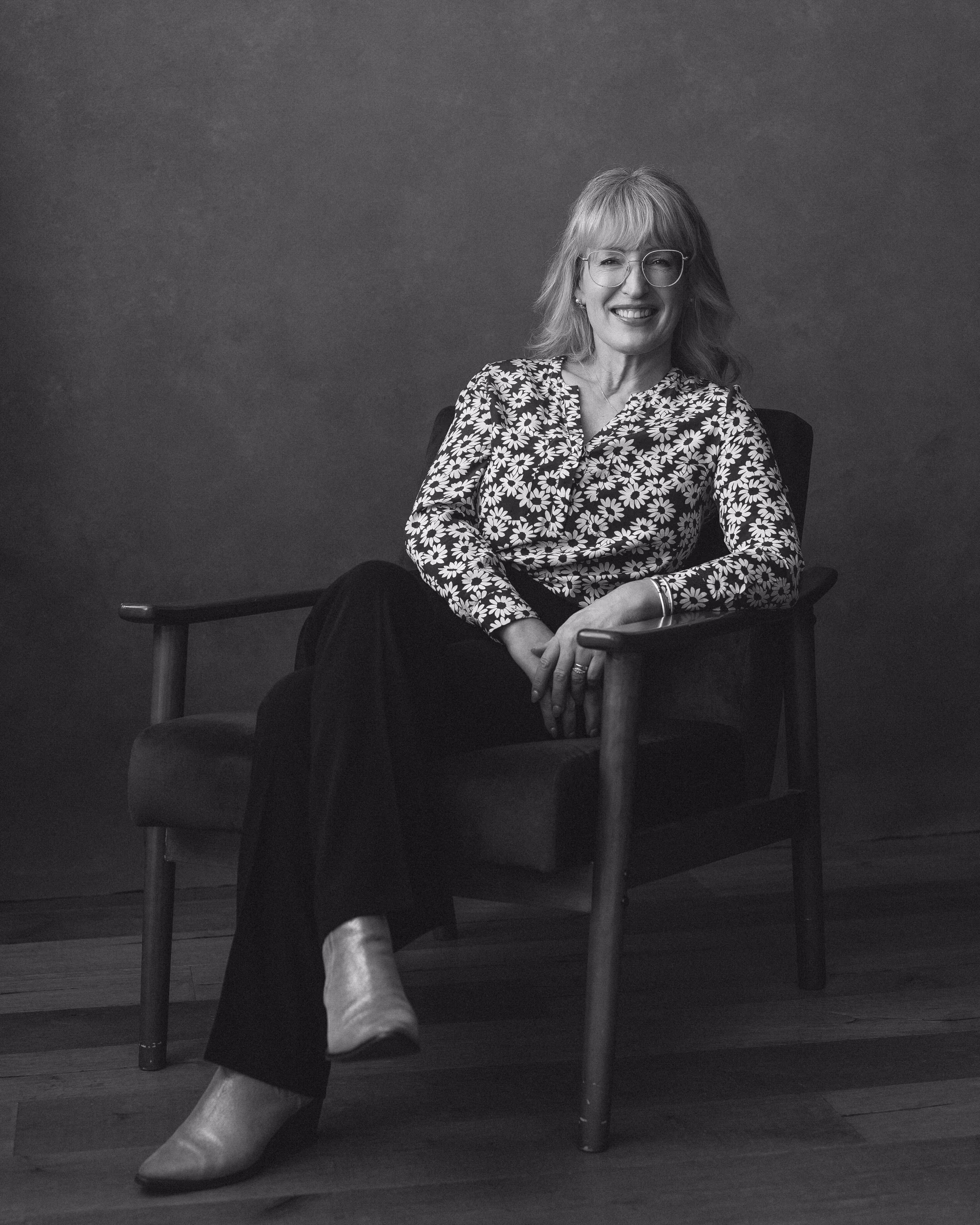 Smiling woman with glasses in a floral top sitting cross-legged on a wooden armchair.