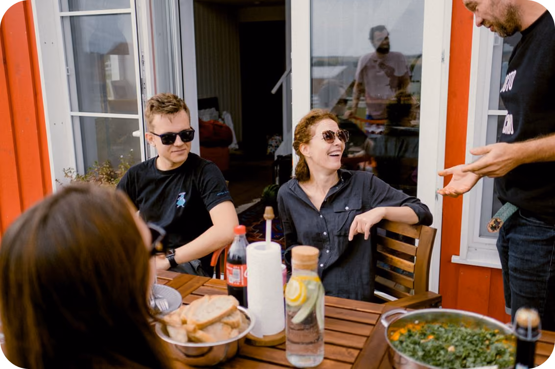 Four people with sunglasses talking and laughing around a wooden table with food and drinks outdoors.