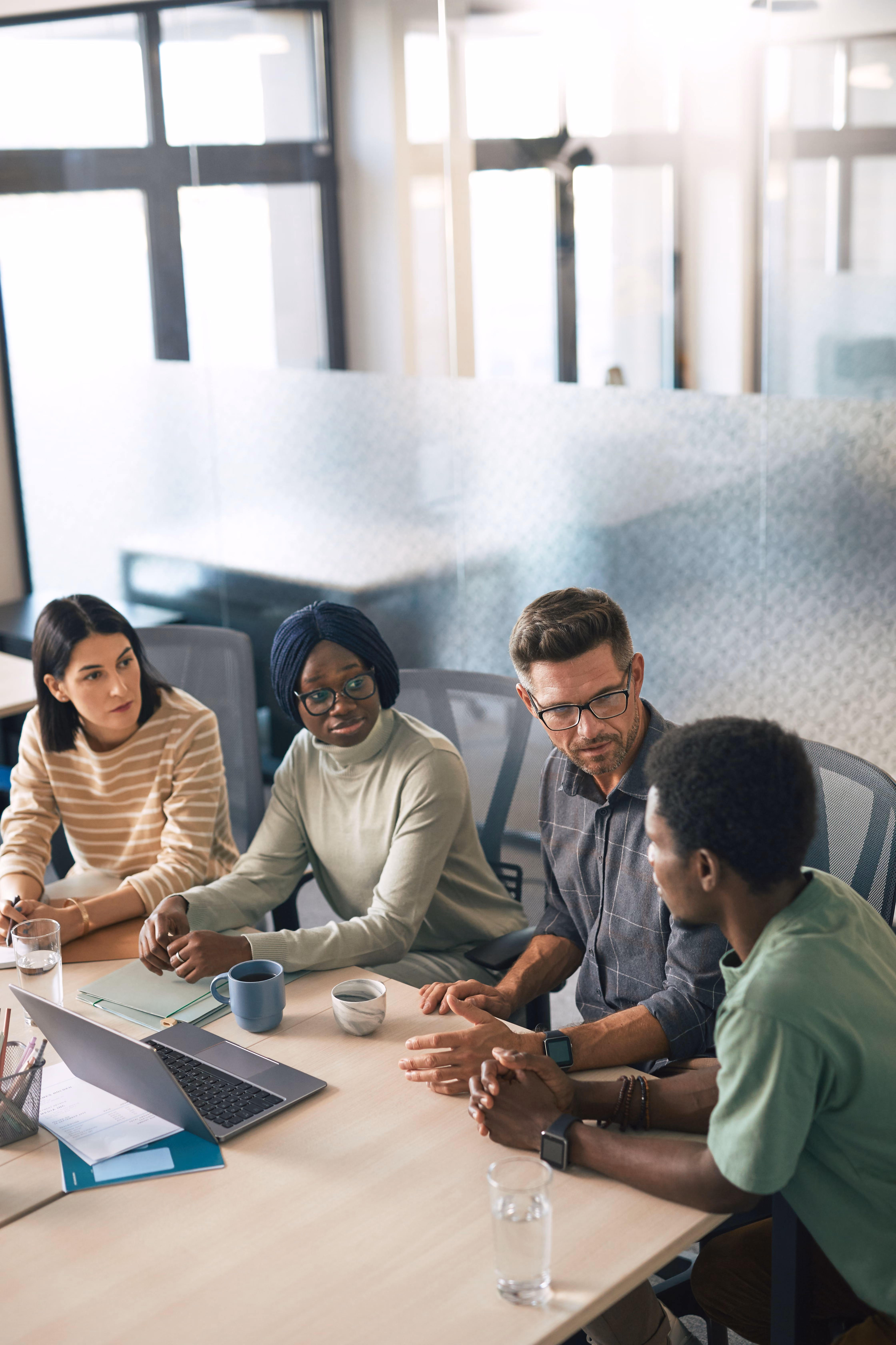 Four diverse coworkers having a meeting around a table with a laptop and cups in a bright office.