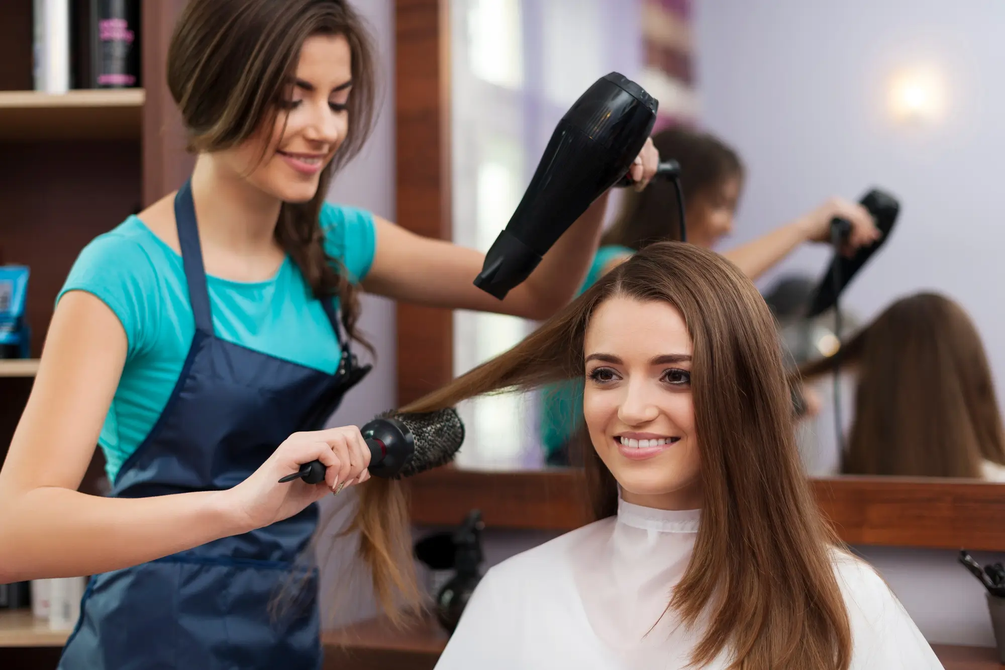 Hairdresser in a teal shirt and dark apron blow-drying and brushing a smiling woman's straight, brown hair in a salon with a mirror reflecting both.