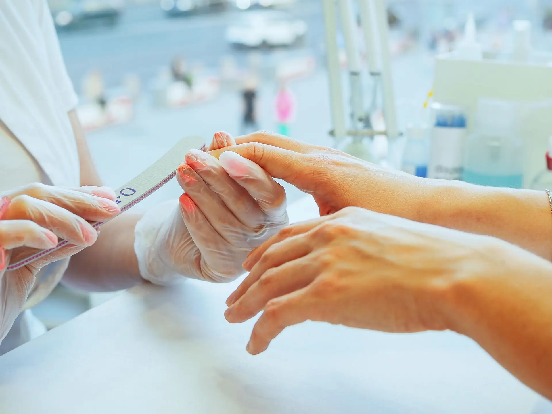 Close-up of a person receiving a manicure; a nail technician wearing disposable gloves is filing the client's nails over a white surface with blurred background of salon items.