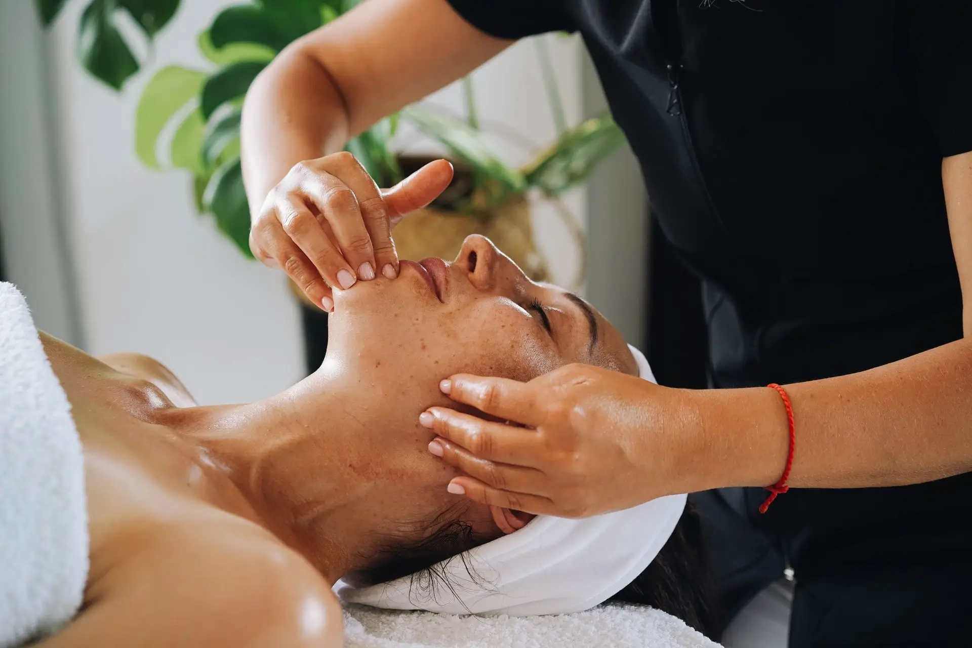 A woman with a white headband lying down with her eyes closed as another person gives her a facial massage, gently touching her chin and jawline.