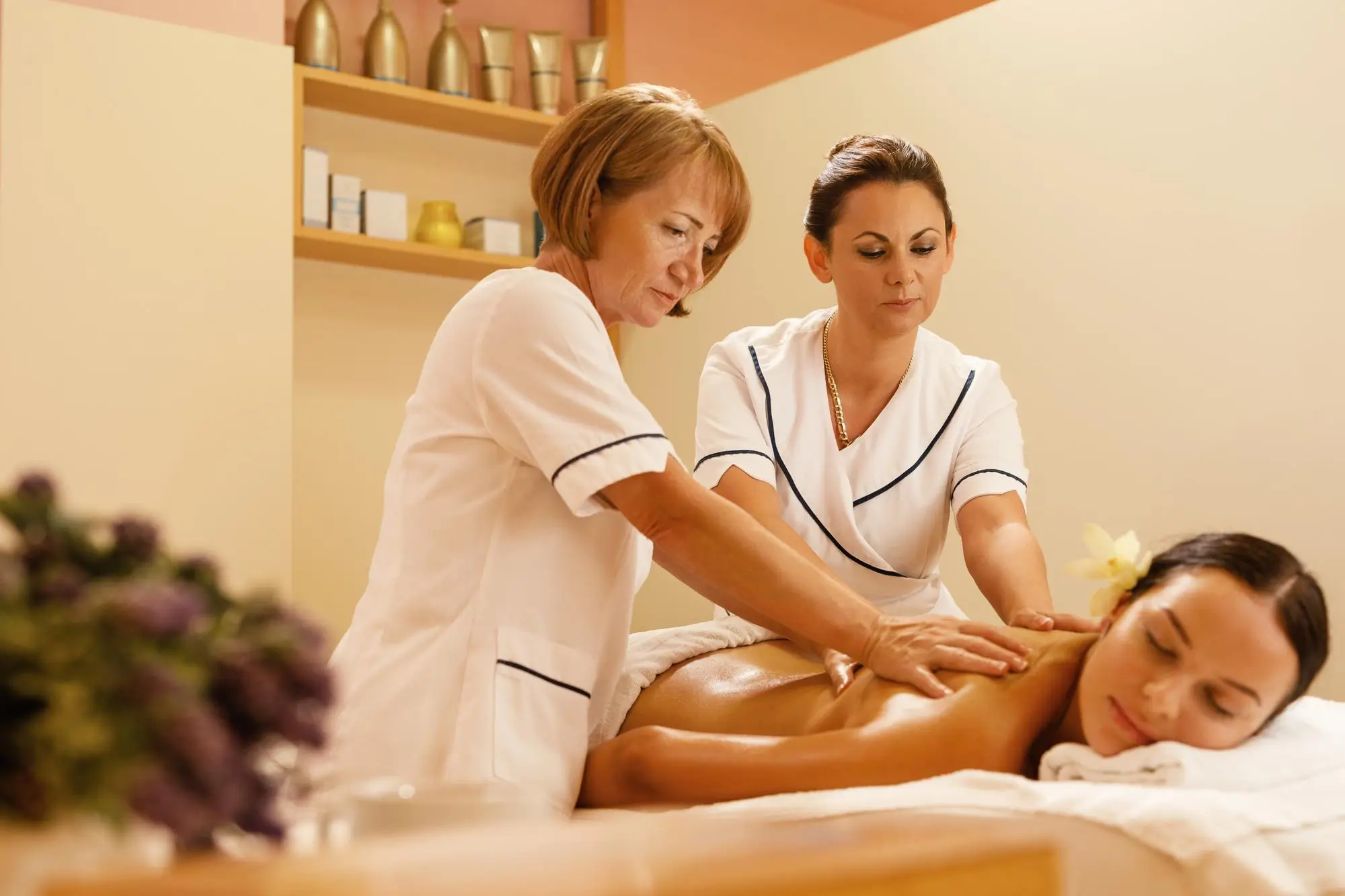 Two spa therapists in white uniforms giving a back massage to a woman lying face down on a massage table with a flower in her hair, in a softly lit spa room.