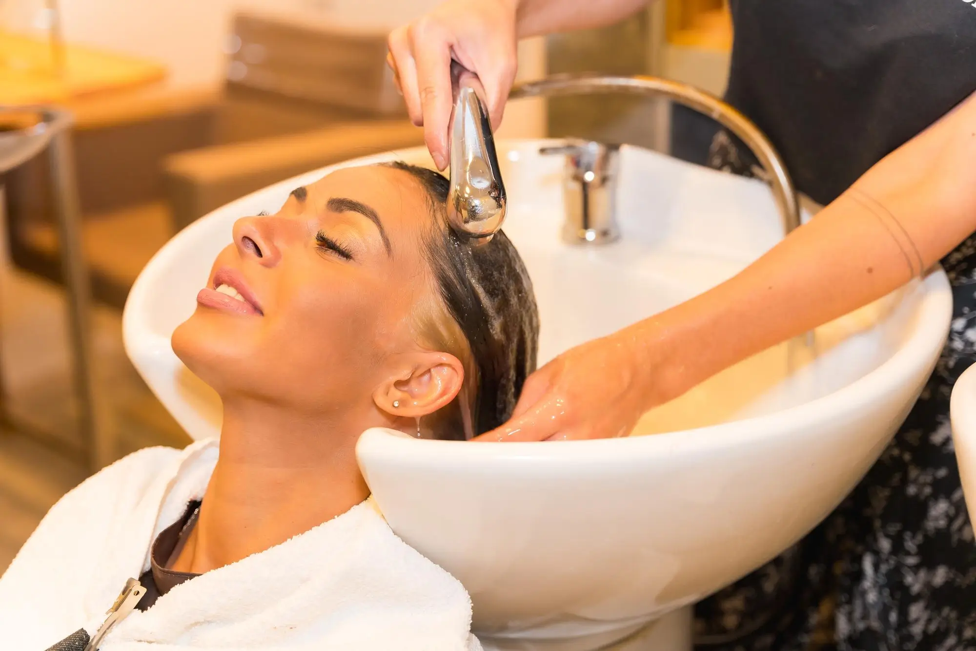 A woman with closed eyes leans back with a white towel around her shoulders as her hair is being washed with water in a salon sink by a person holding a shower head.