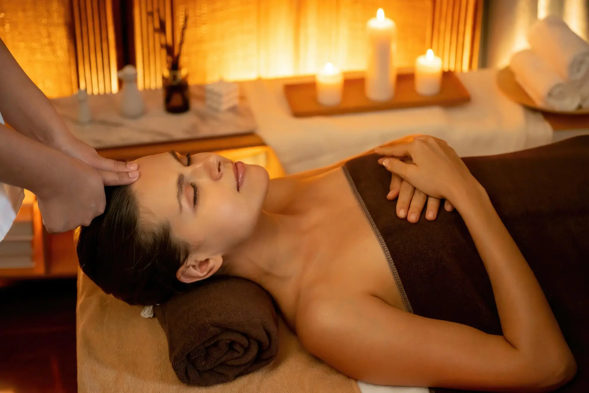 Woman lying on a massage table with eyes closed and a brown towel covering her body, receiving a forehead massage in a softly lit spa room with candles and rolled towels in the background.