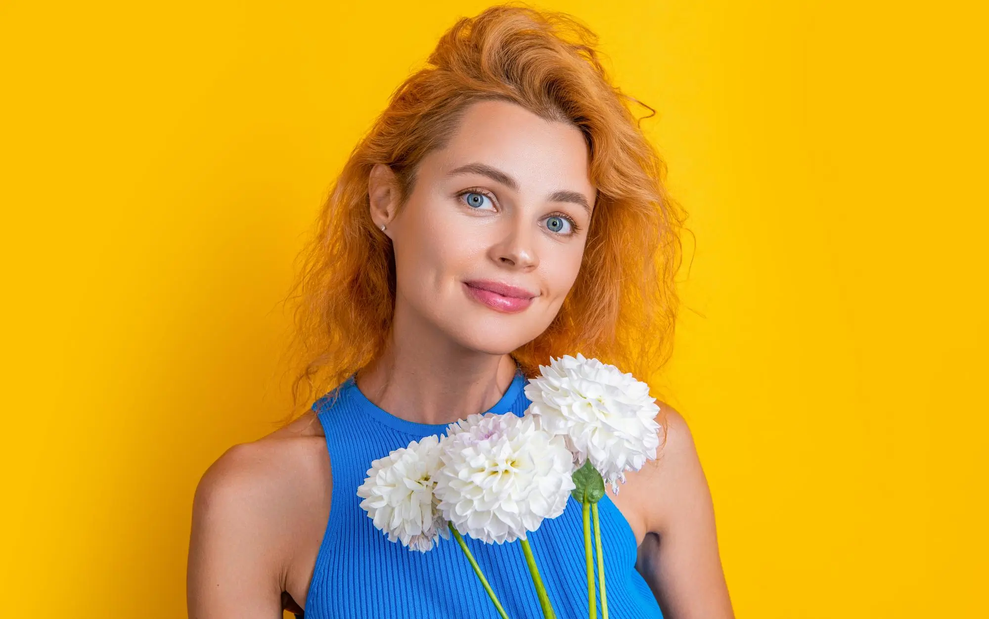 Smiling woman with light skin and wavy red hair wearing a blue sleeveless top holding three white flowers in front of a bright yellow background.