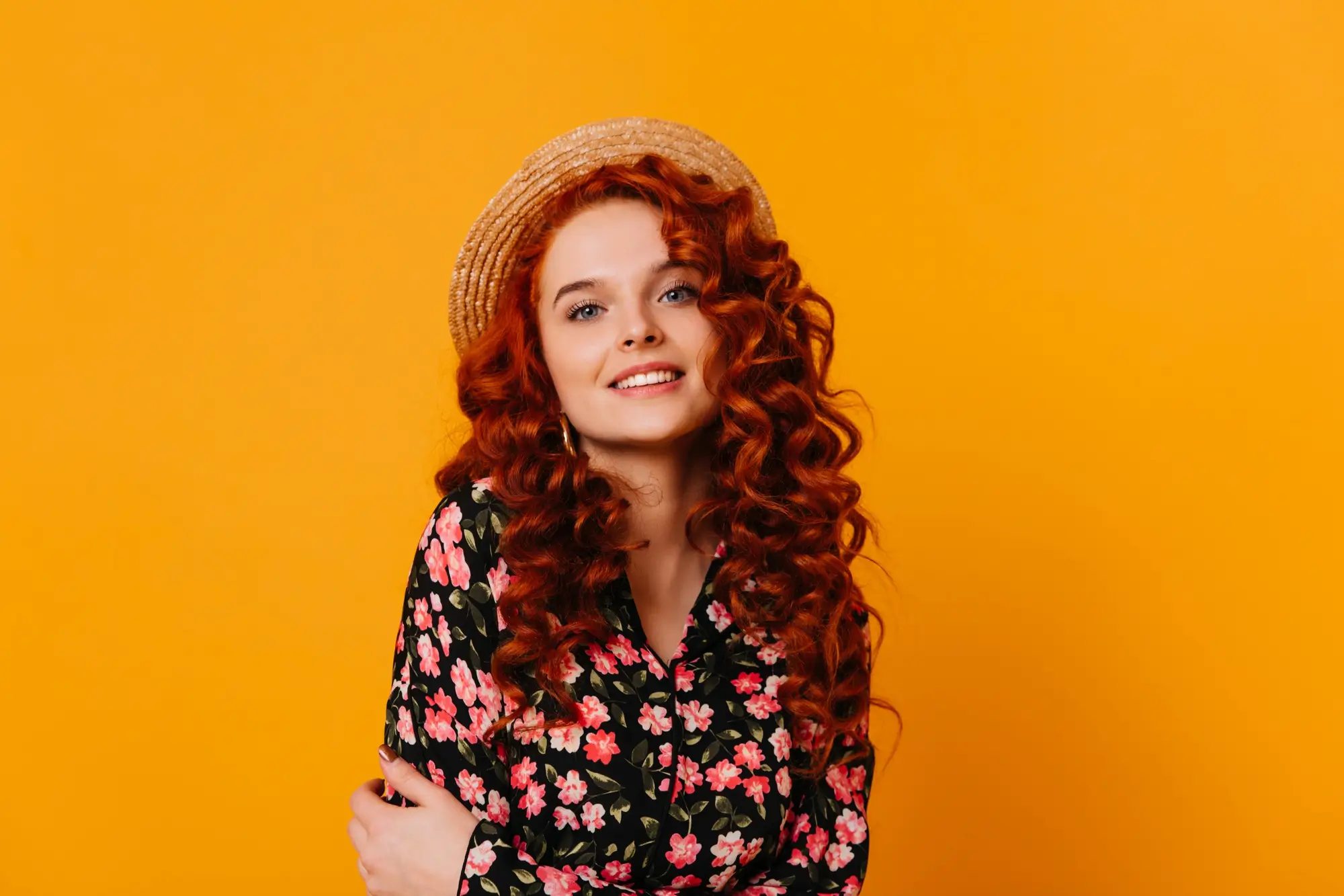 Young woman with long curly red hair wearing a straw hat and a black floral shirt, smiling against an orange background.