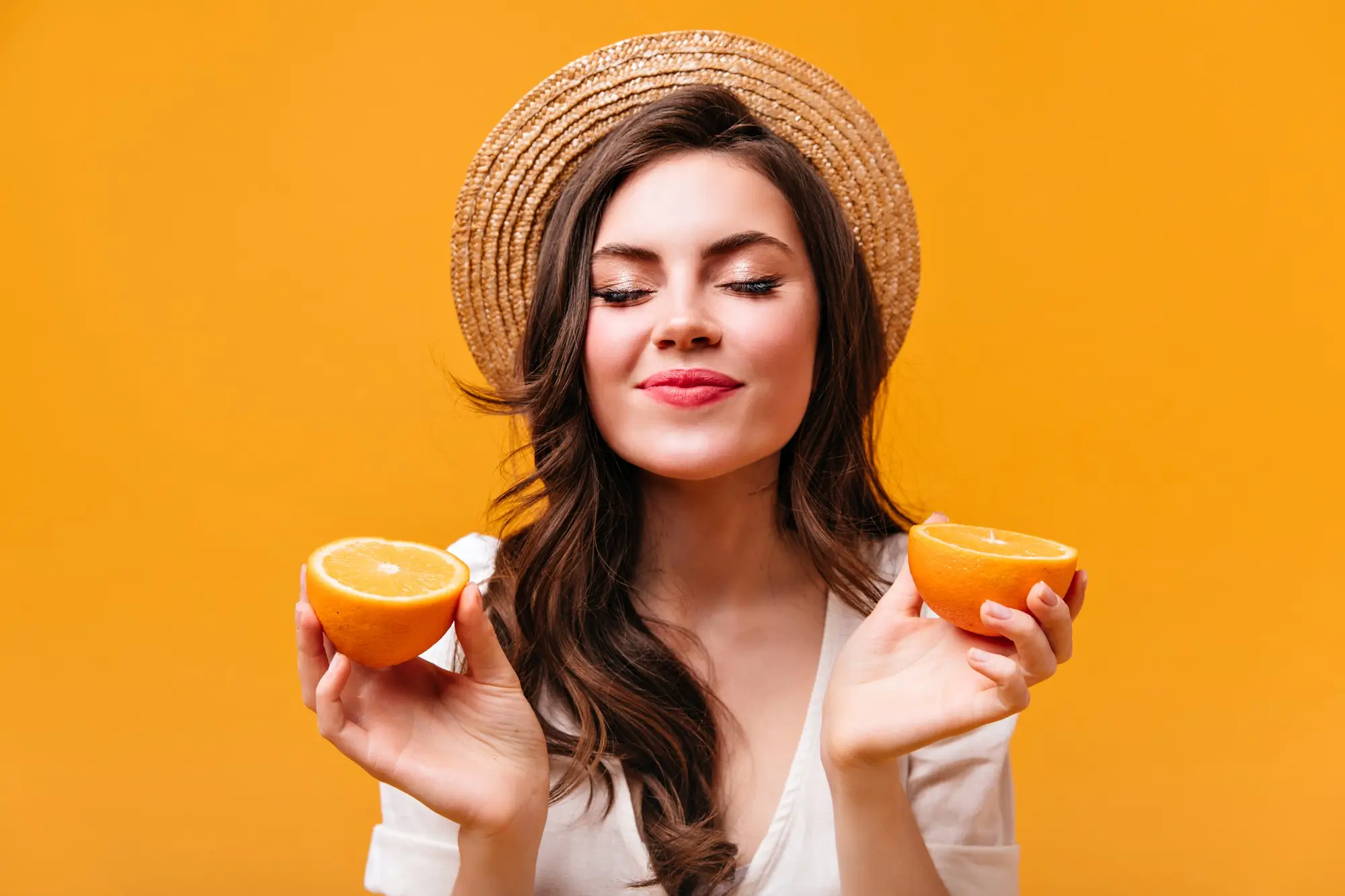 Young woman with long brown hair wearing a straw hat and white top holding two halves of an orange with eyes closed and a slight smile against an orange background.