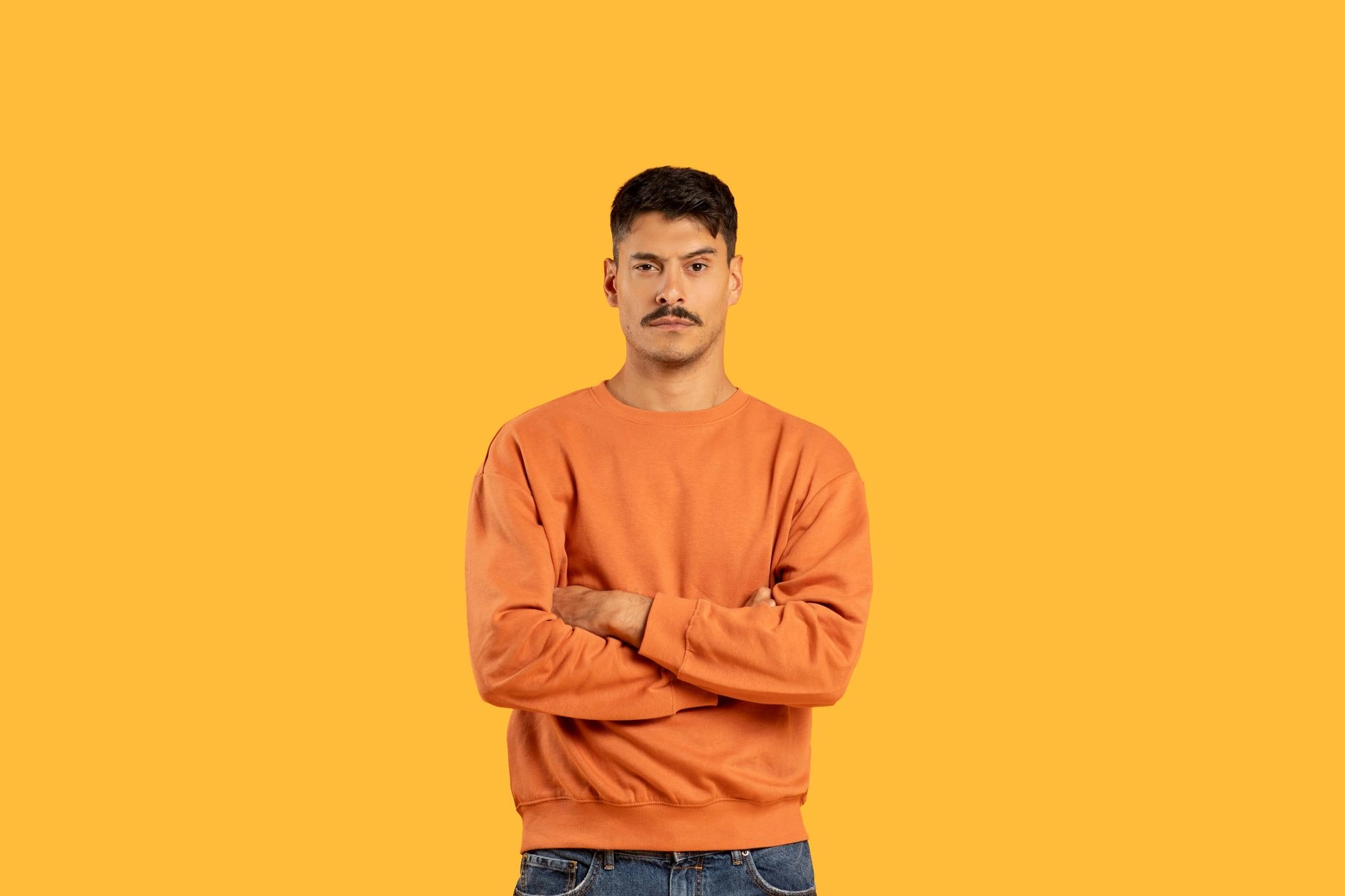 Man with dark hair and mustache standing against a solid yellow background, wearing an orange sweatshirt and blue jeans, arms crossed with a serious expression.