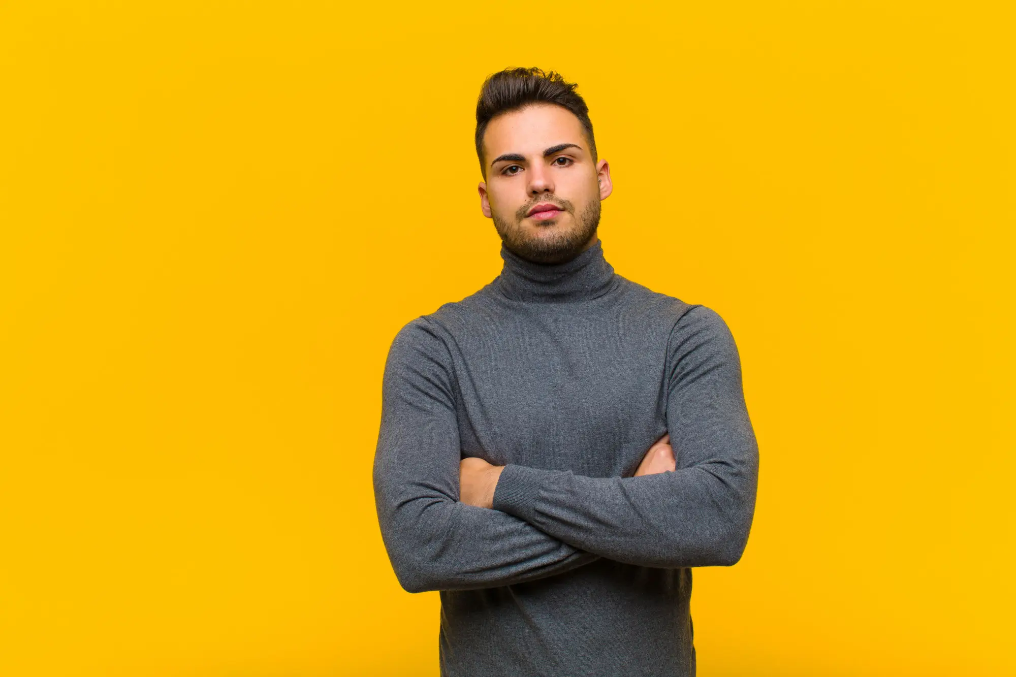 Young man with dark hair and beard wearing a gray turtleneck sweater, standing with arms crossed against a solid bright yellow background.
