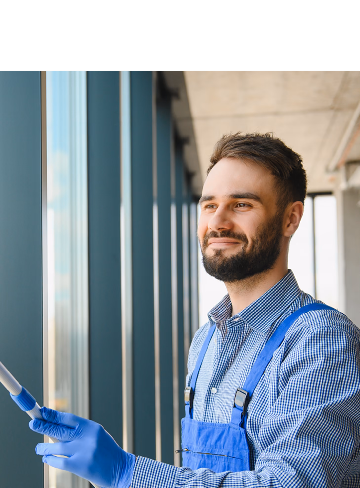 Smiling man wearing blue gloves and a blue apron, holding a cleaning tool while looking out a large window in a modern building.