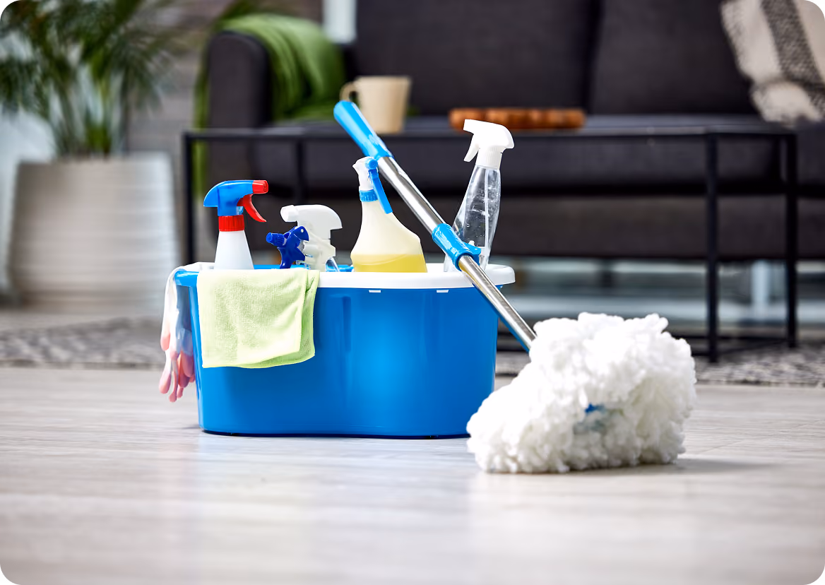 Blue cleaning caddy with spray bottles, gloves, and a cloth, next to a mop with a white head on a tiled floor in a living room.