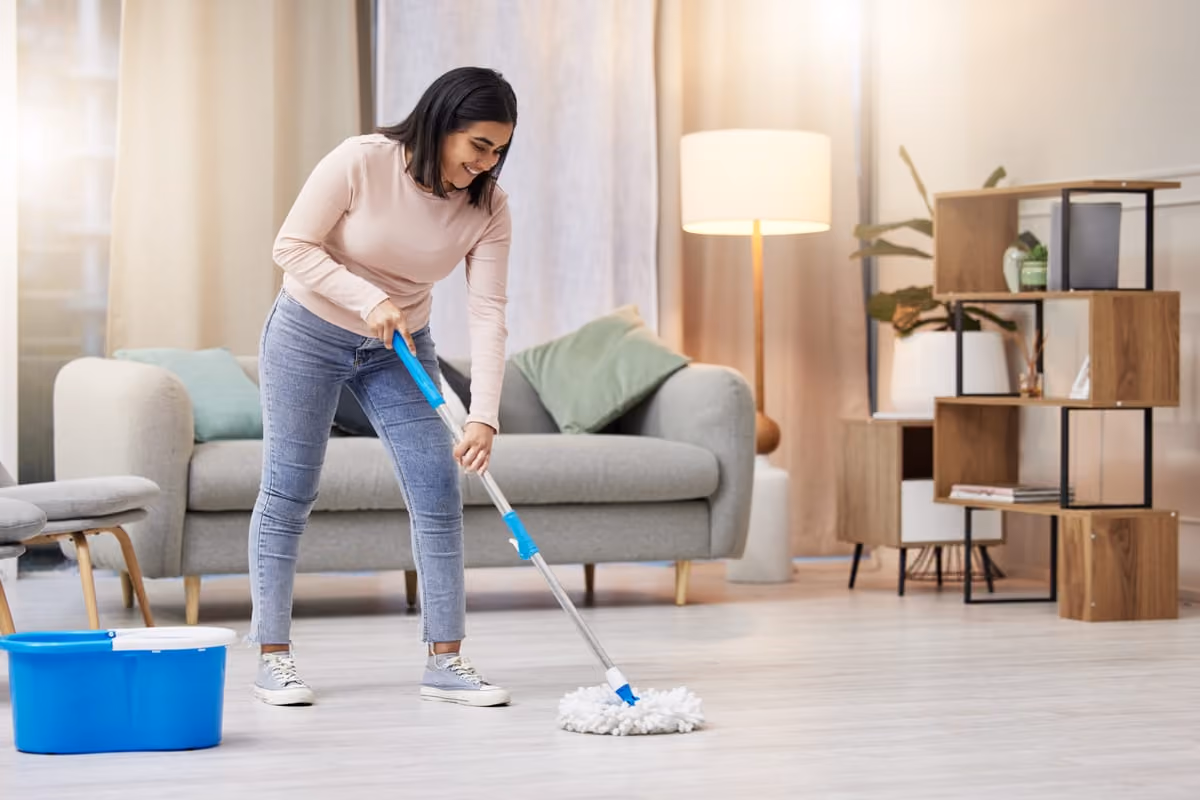 Woman mopping a light-colored floor in a cozy living room with a gray sofa, wooden shelving, floor lamp, and plants.