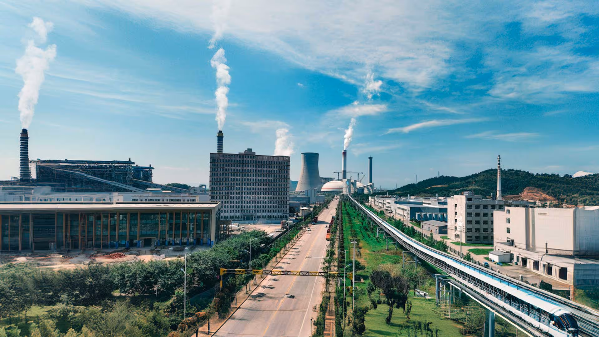 A modern industrial complex with multiple buildings, chimneys emitting white smoke, and a train on an elevated track running parallel to a road lined with trees.