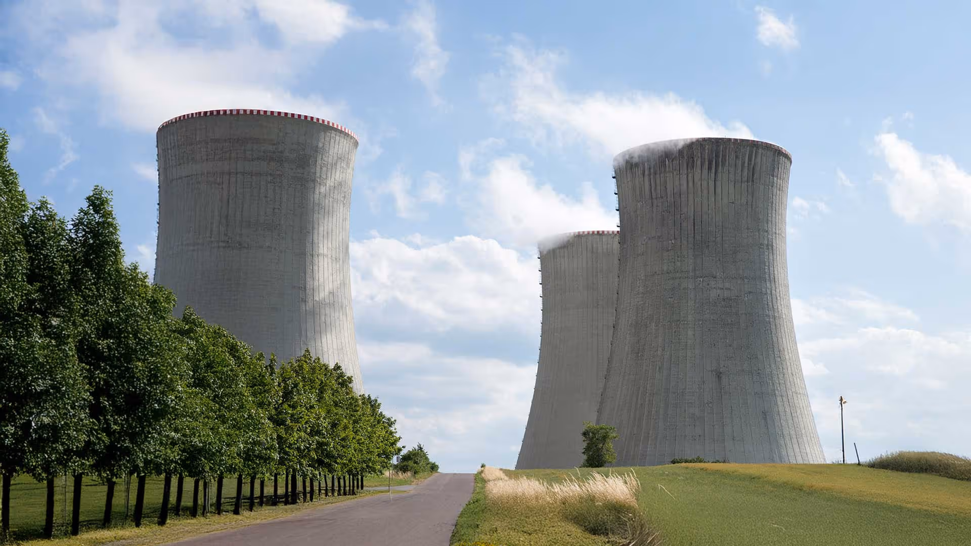 Three large cooling towers emitting steam at a power plant with a road and trees in the foreground under a blue sky with clouds.