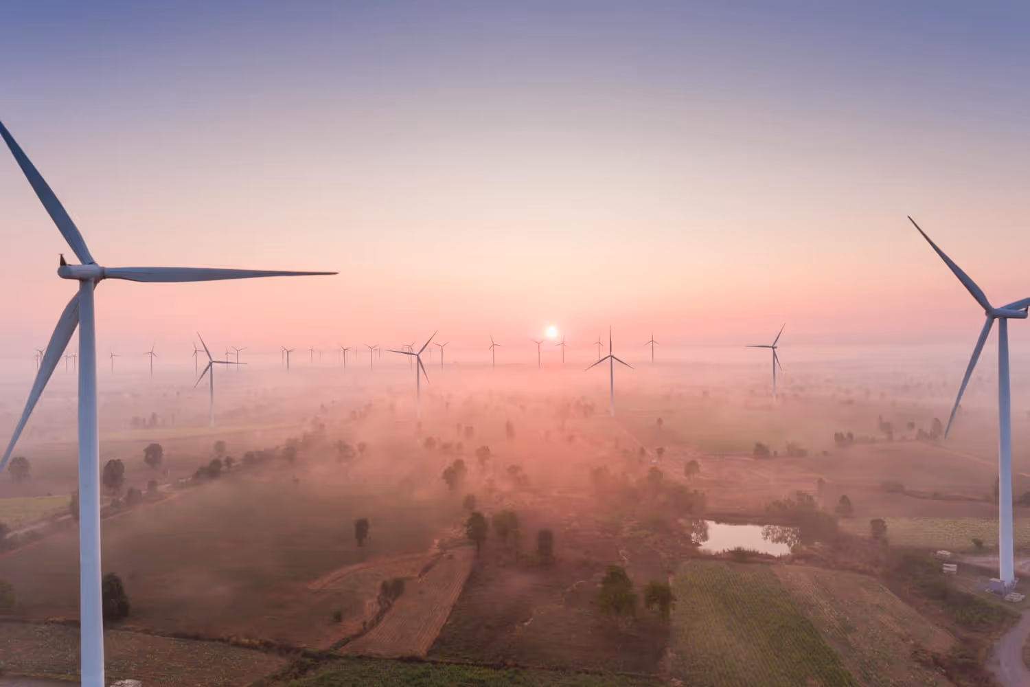 Wind turbines spread across a rural landscape at sunrise with a pink and purple sky and mist over the fields.