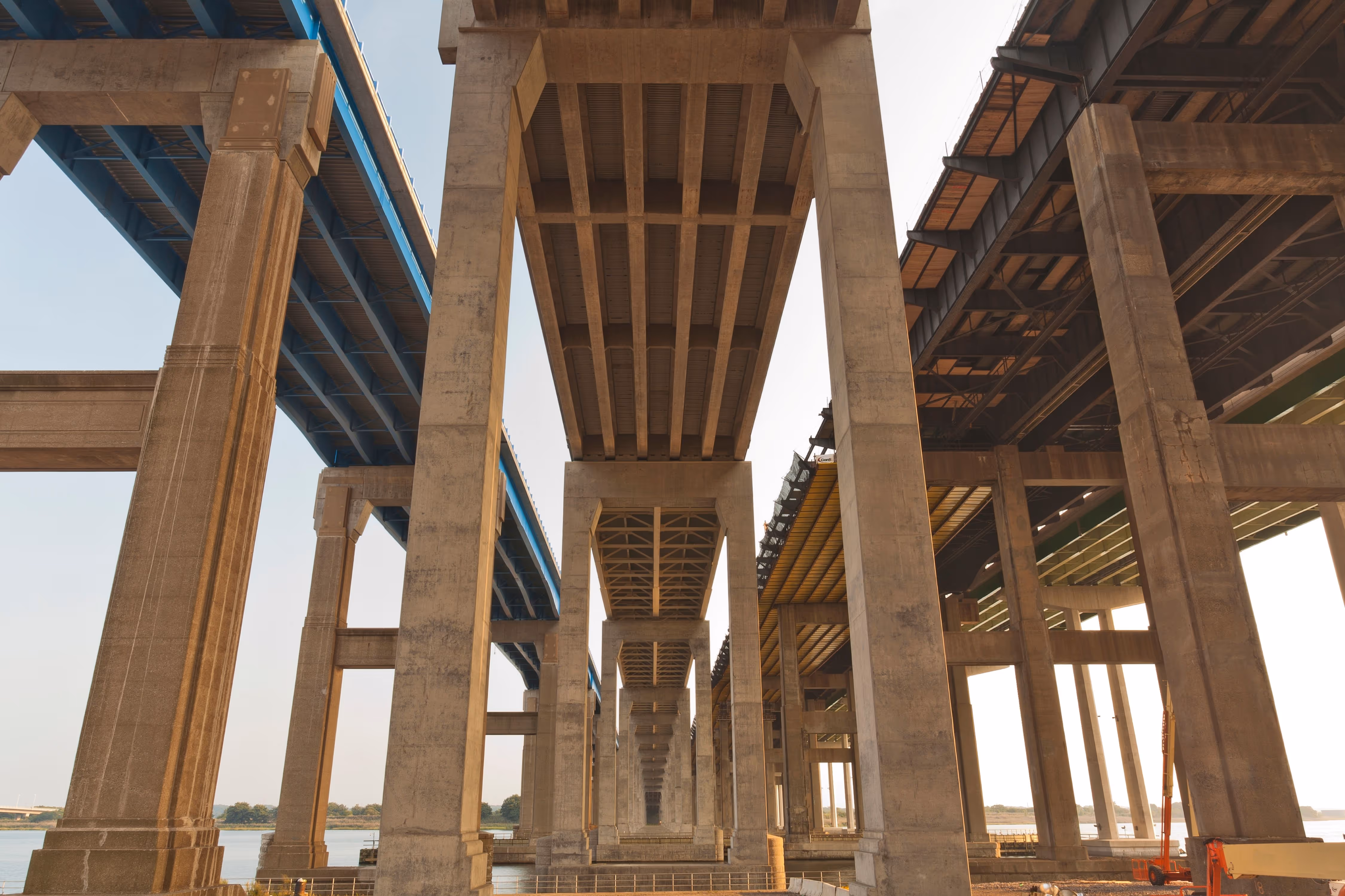 Underside view of multiple large concrete bridges with pillars over a river during daytime.