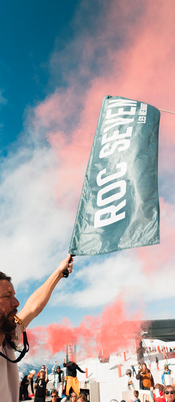 Un homme avec une sifflet dans la bouche agite un drapeau vert avec l'inscription blanche « ROC SEVEN » sous un ciel bleu avec des fumées roses, avec une foule de personnes en arrière-plan sur une pente enneigée.