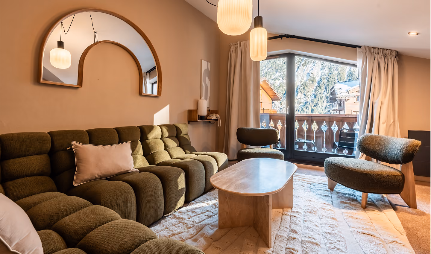 Salon moderne avec un canapé vert olive à coussins rembourrés, deux fauteuils assortis, une table basse en bois clair, un grand miroir décoratif au mur, des suspensions lumineuses blanches et une porte-fenêtre donnant sur un balcon en bois avec vue sur des arbres enneigés.