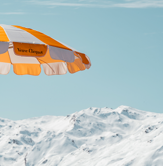 Parasol orange et blanc Veuve Clicquot au premier plan avec des montagnes enneigées en arrière-plan sous un ciel bleu clair.