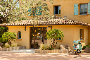 Façade d'une maison ocre avec un toit en tuiles, une porte vitrée entourée de petits arbres et un scooter bleu garé à droite.