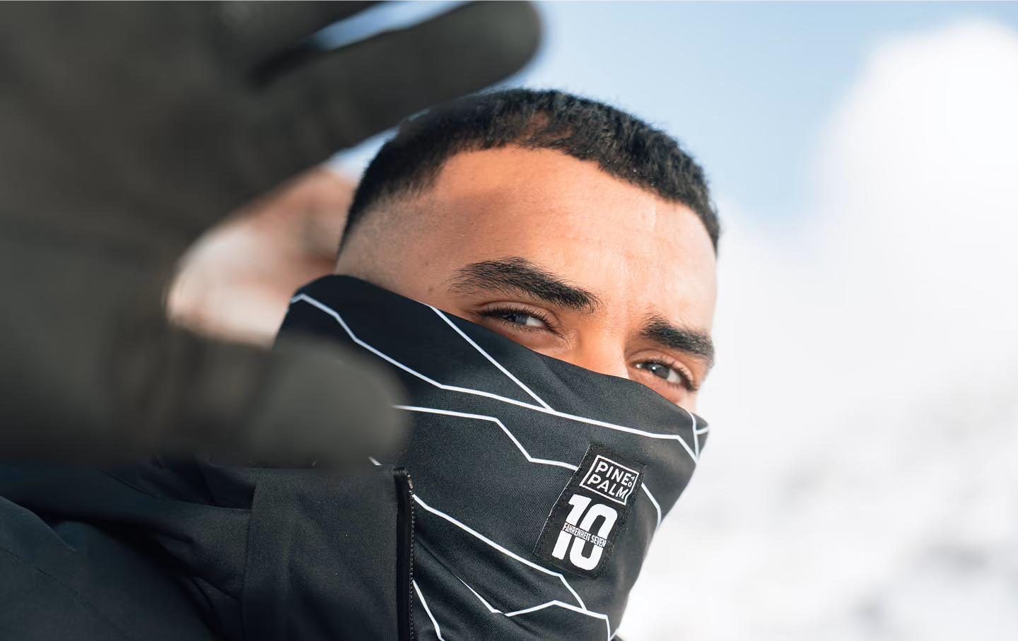 Homme avec des cheveux courts portant un masque noir couvrant sa bouche et son nez, avec un gant noir approchant la caméra, fond flou de ciel clair.