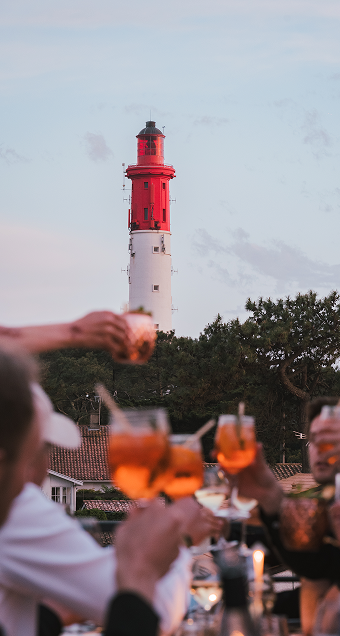 Un phare rouge et blanc se dresse derrière des arbres et des toits au coucher du soleil, tandis que des personnes lèvent des verres avec une boisson orange en premier plan.