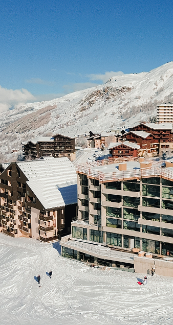 Vue sur des immeubles et chalets en bois dans une station de ski enneigée, avec une montagne enneigée en arrière-plan et un ciel bleu clair.