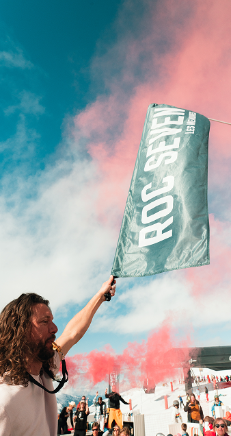 Un homme avec une barbe et des cheveux longs agitant un drapeau marqué ROC SEVEN devant une foule en plein air sous un ciel bleu avec de la fumée rouge en arrière-plan.