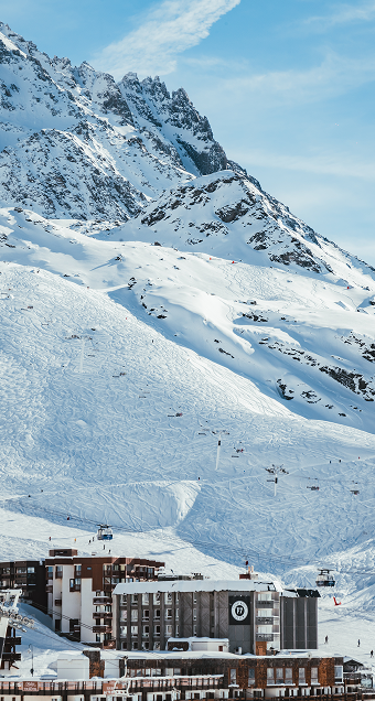 Station de ski au pied d'une montagne enneigée avec des pistes de ski marquées, des skieurs et des remontées mécaniques en arrière-plan sous un ciel bleu avec quelques nuages.