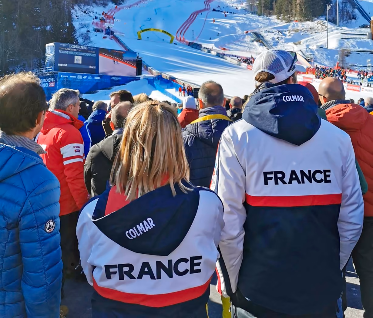 Des spectateurs en veste FRANCE regardant une piste de ski enneigée pendant une compétition de ski, avec une foule et des panneaux visibles en arrière-plan.