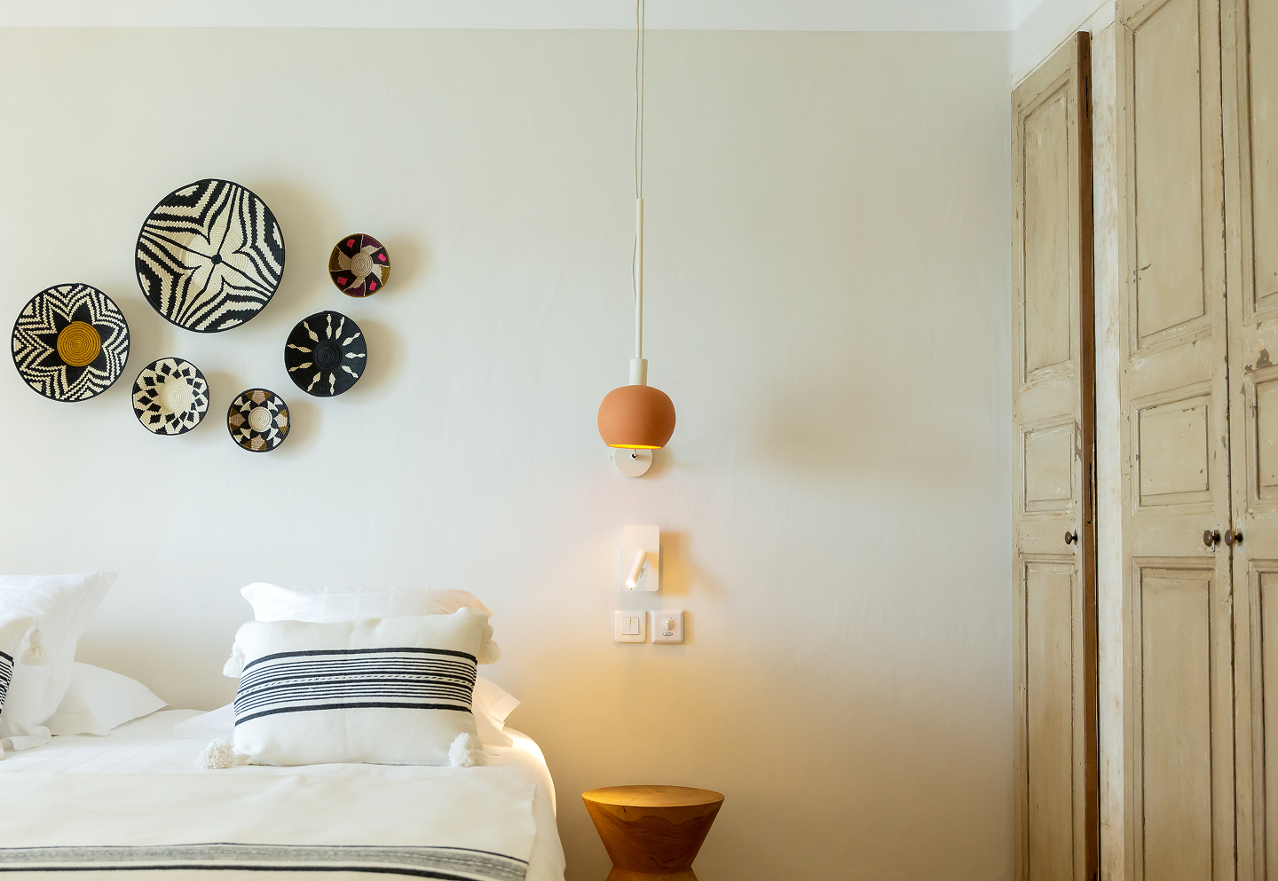 Chambre minimaliste avec lit double à linge blanc, coussin rayé noir et blanc, murs clairs décorés de six paniers muraux tissés aux motifs noirs, blancs et marron, lampe suspendue orange, tabouret en bois et armoire en bois clair usé.