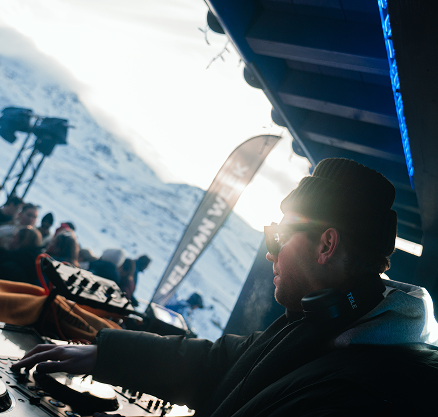 Un DJ portant des lunettes de soleil, un bonnet et un casque autour du cou, joue de la musique en extérieur avec des montagnes enneigées en arrière-plan.