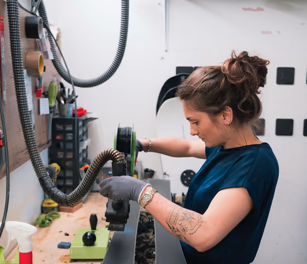 Une femme avec un tatouage sur le bras travaille sur un équipement dans un atelier, tenant une pièce avec des gants et concentrée sur la tâche.