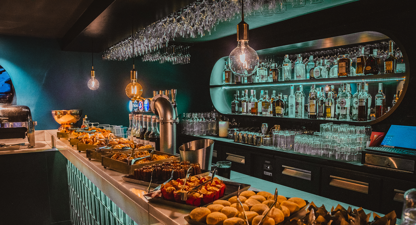 Bar élégant avec un comptoir où sont disposés plusieurs plateaux de petits pains et pâtisseries variées, éclairage par trois ampoules suspendues, étagères en arrière-plan remplies de bouteilles d'alcool et de verres, ainsi qu'un ordinateur portable ouvert.