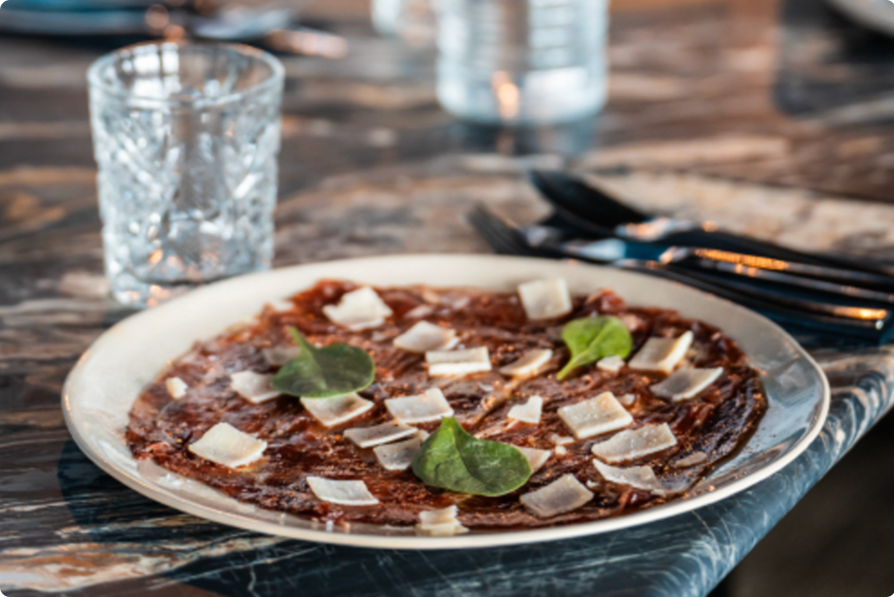 Assiette de viande hachée finement tranchée garnie de copeaux de parmesan et de feuilles d'épinard, posée sur une table en marbre avec un verre vide et des couverts à côté.