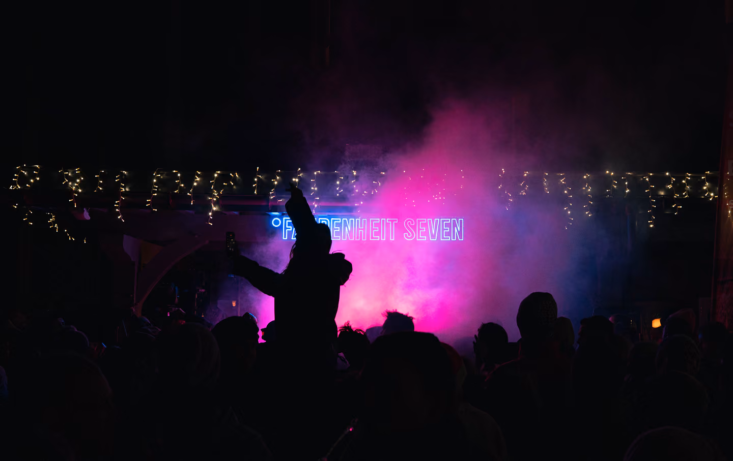 Silhouettes of a crowd at a night event with one person holding a phone up amid pink and purple fog, festive string lights, and a neon sign reading 'Fahrenheit Seven'.