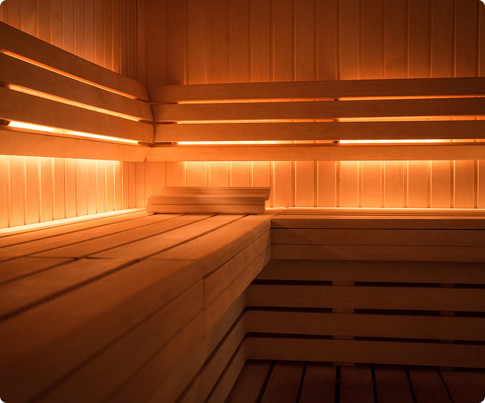 Intérieur d'un sauna en bois aux lumières chaudes intégrées sous les bancs et panneaux muraux, avec un repose-tête en bois posé sur le banc.