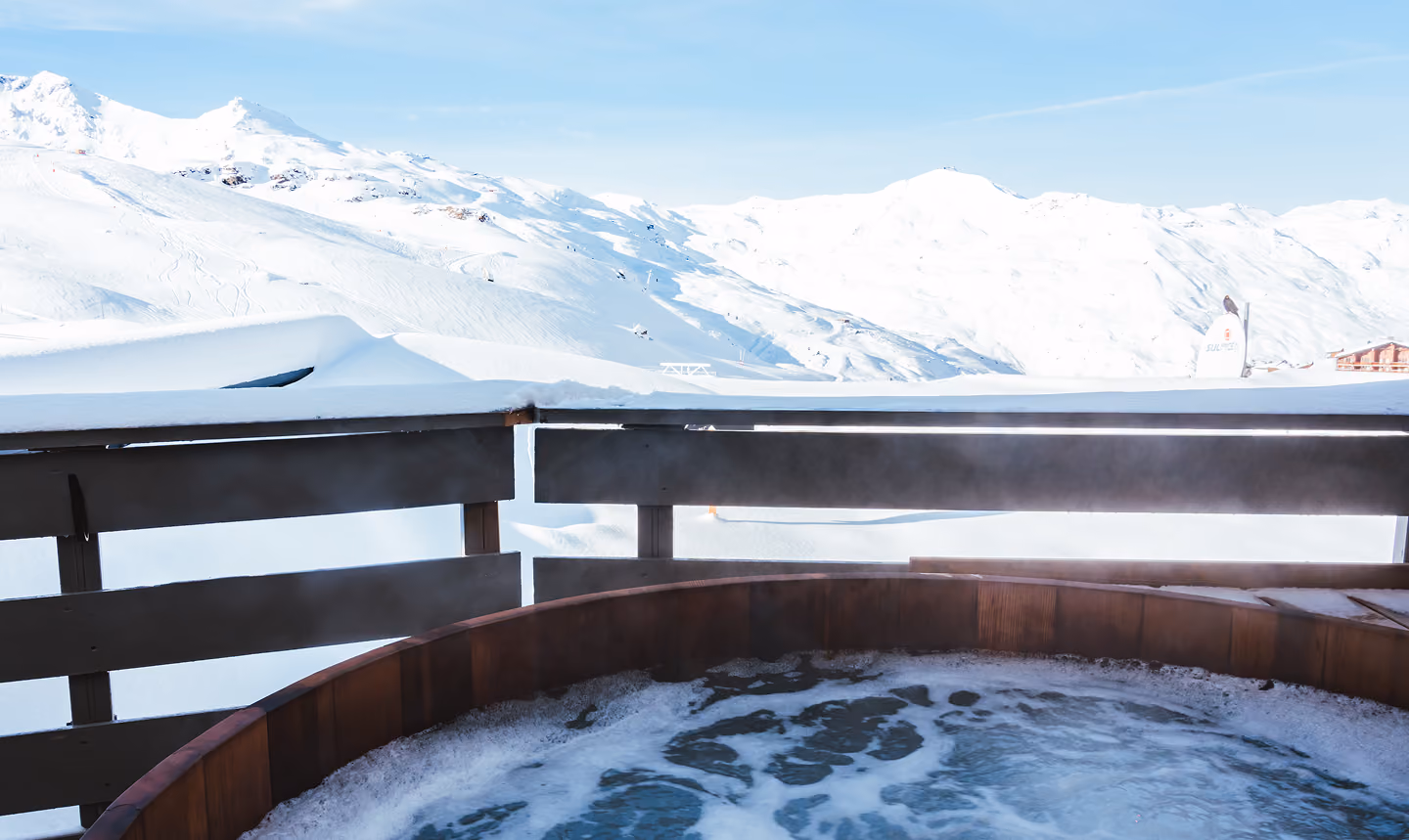 Baignoire extérieure en bois avec eau bouillonnante sur un balcon offrant une vue sur des montagnes enneigées sous un ciel bleu clair.