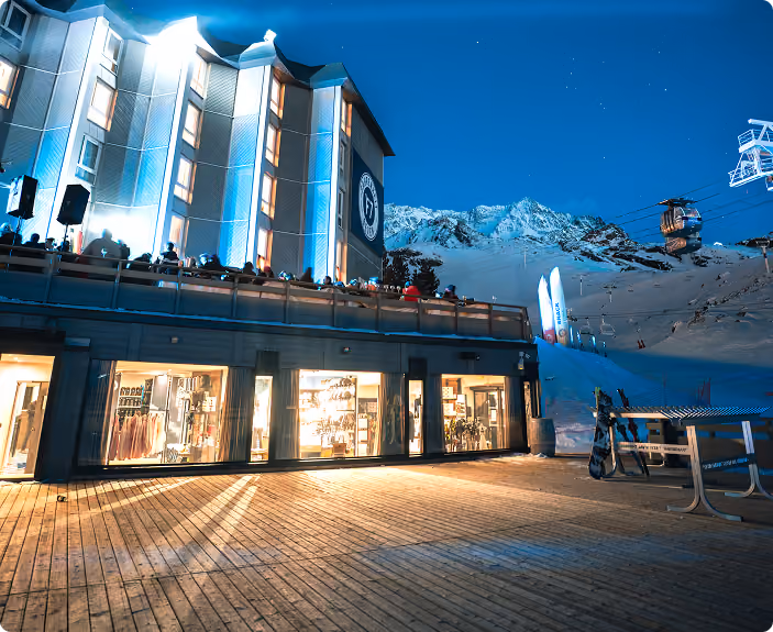 Vue de nuit d'un bâtiment éclairé avec une terrasse peuplée de personnes, devant une montagne enneigée avec une télécabine en fonctionnement sous un ciel étoilé.