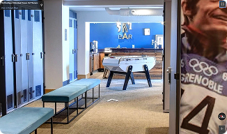 Vue d'un couloir intérieur menant à un bar étiqueté "SKI BAR" avec un baby-foot au centre, des bancs à gauche et des casiers le long du mur, et une affiche partielle d'un athlète portant un bonnet et un maillot avec le mot "GRENOBLE" visible à droite.