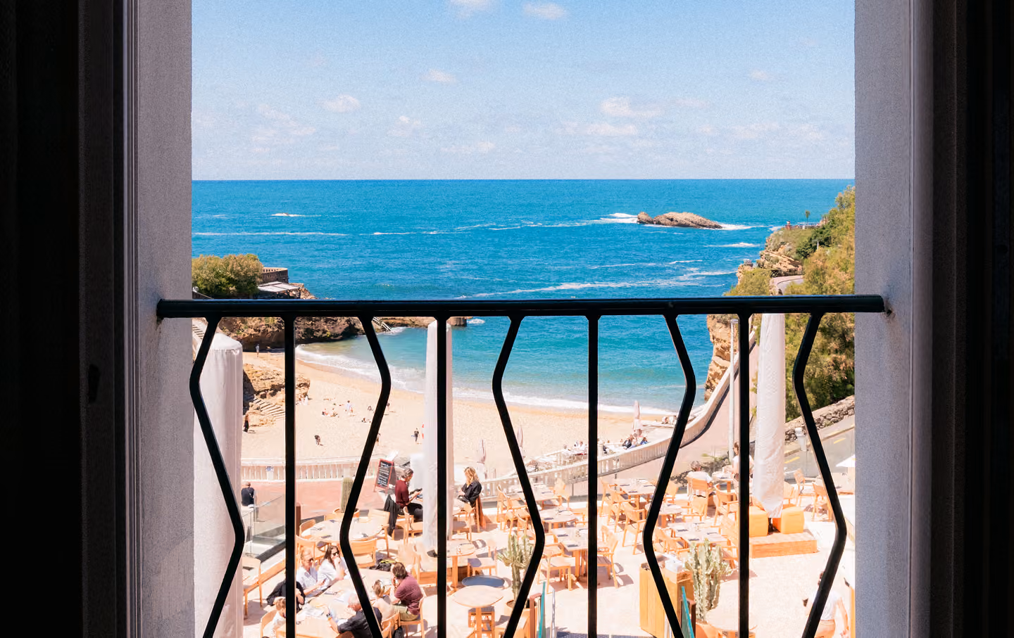 Vue depuis un balcon avec rambarde noire sur une plage ensoleillée, des gens sur le sable et une mer bleue calme sous un ciel légèrement nuageux.