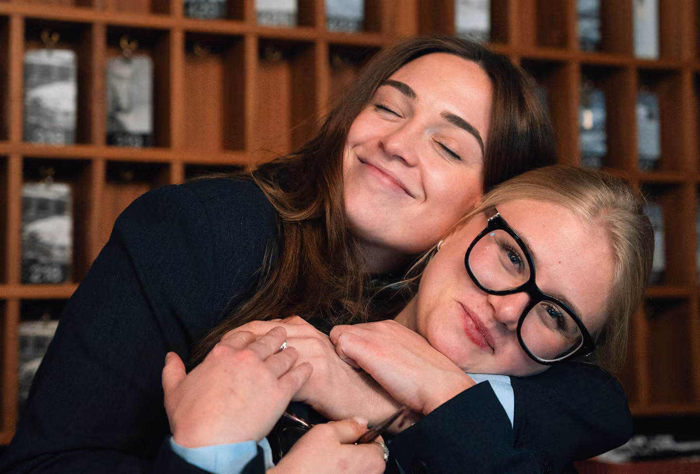Deux femmes qui s'enlacent affectueusement, l'une avec les yeux fermés et un sourire, l'autre portant des lunettes noires et posant la tête sur l'épaule de la première.