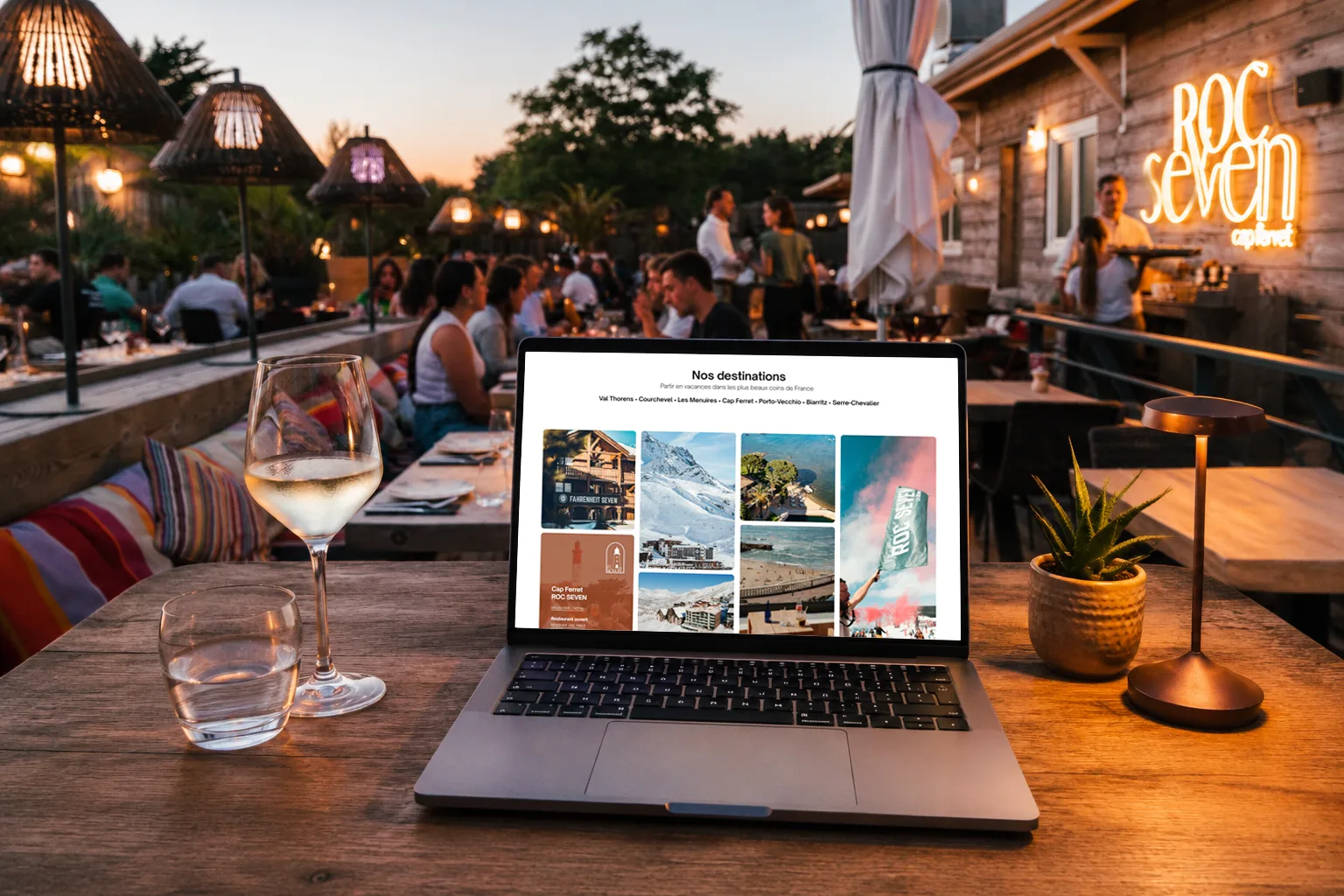 Un ordinateur portable ouvert affichant une page de destinations de vacances en France, posé sur une table en bois avec un verre de vin blanc, un verre d'eau, une plante en pot et une lampe. En arrière-plan, un restaurant en plein air avec des clients et des lumières tamisées au crépuscule.