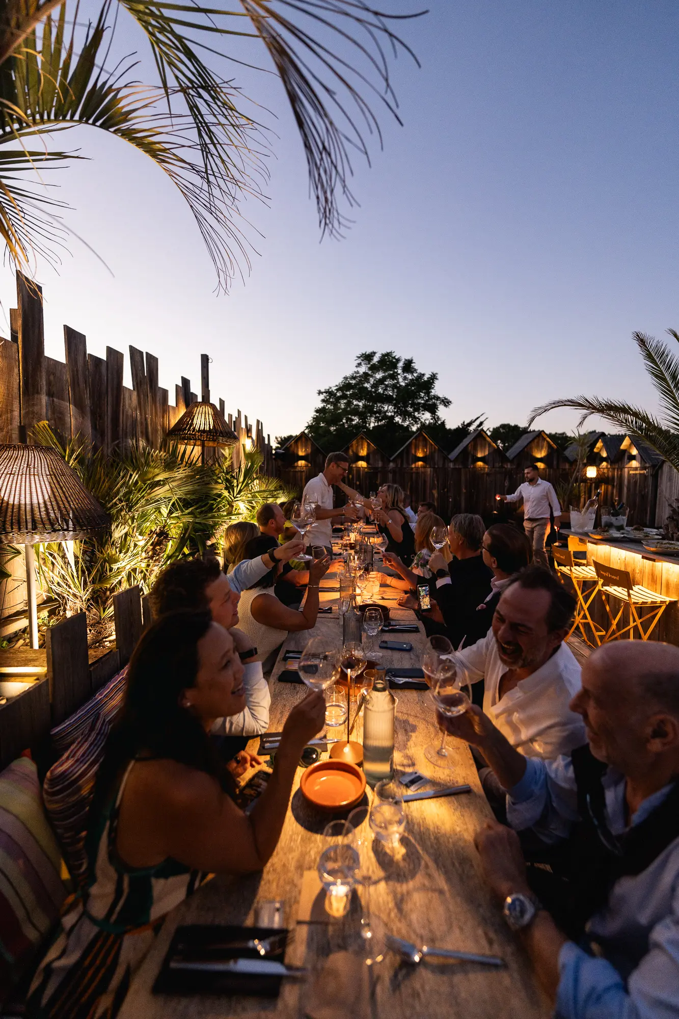 Groupe de personnes assises à une longue table extérieure en bois, levant leurs verres pour un toast au crépuscule avec des lumières chaudes et des palmiers autour.