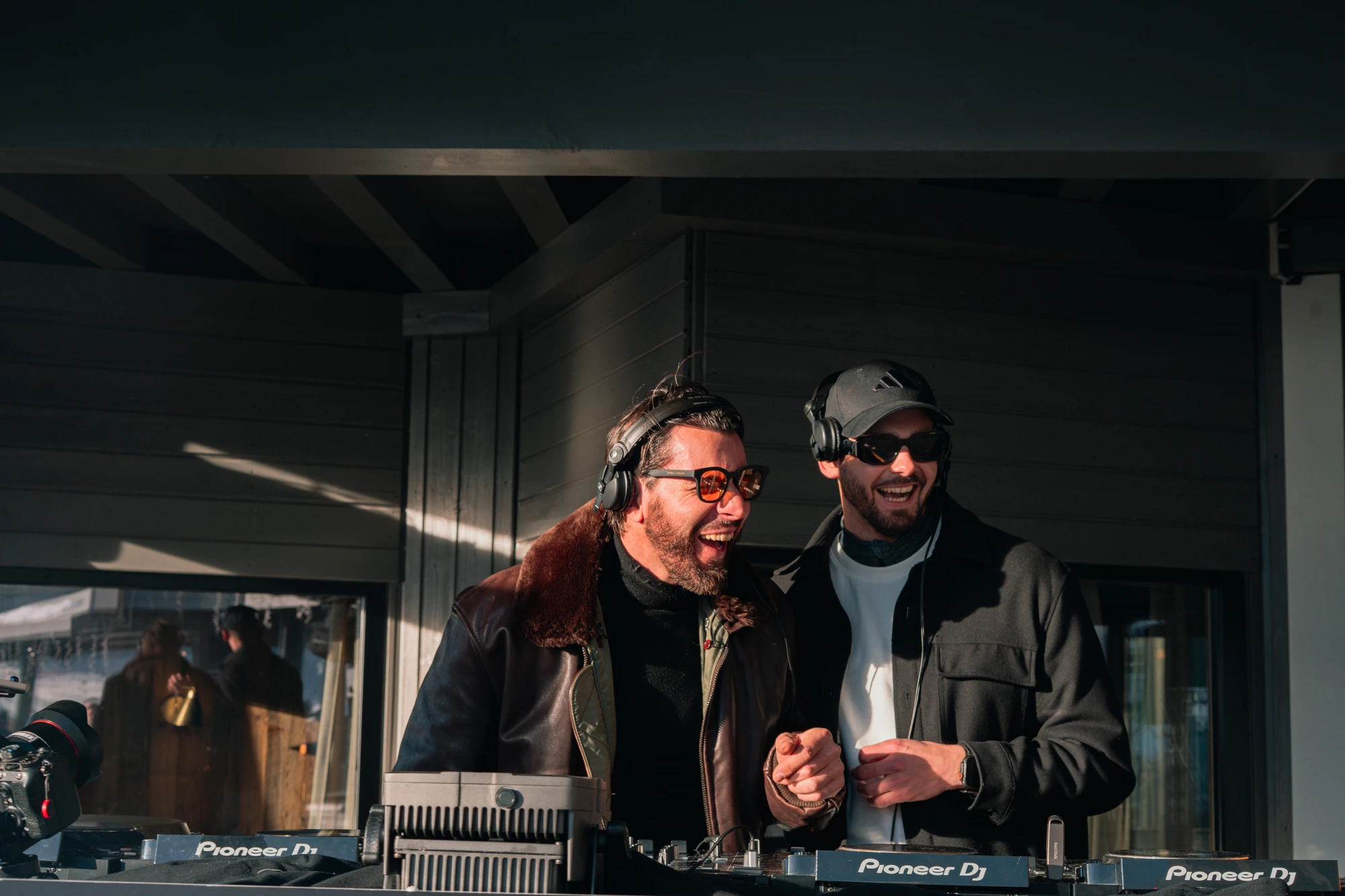 Deux DJs portant des casques et des lunettes de soleil sourient devant leur équipement DJ Pioneer sur une scène extérieure.