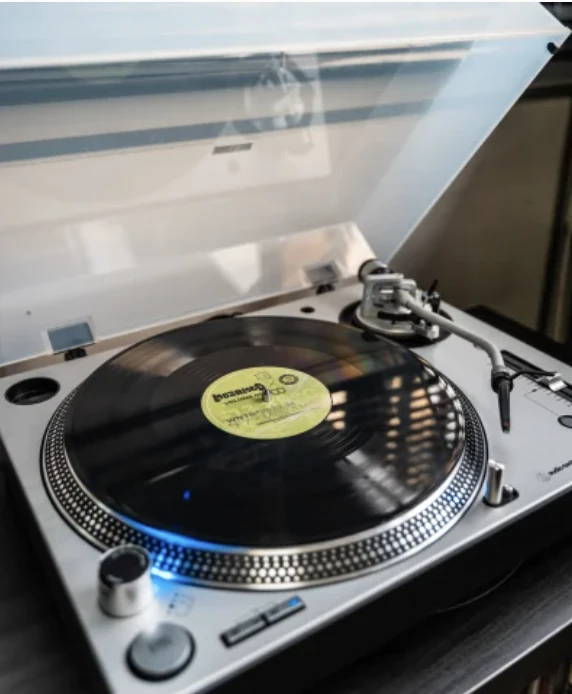 Platine vinyle argentée en fonctionnement avec un disque noir et un label jaune au centre, sous un couvercle transparent légèrement ouvert.