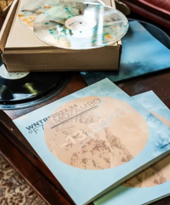 Une table en bois avec plusieurs vinyles, un vinyle transparent avec des taches de couleurs, un vinyle noir, et des brochures bleu clair avec un cercle orange portant les mots 'WNTR 2023-24 MOZAMBO'.