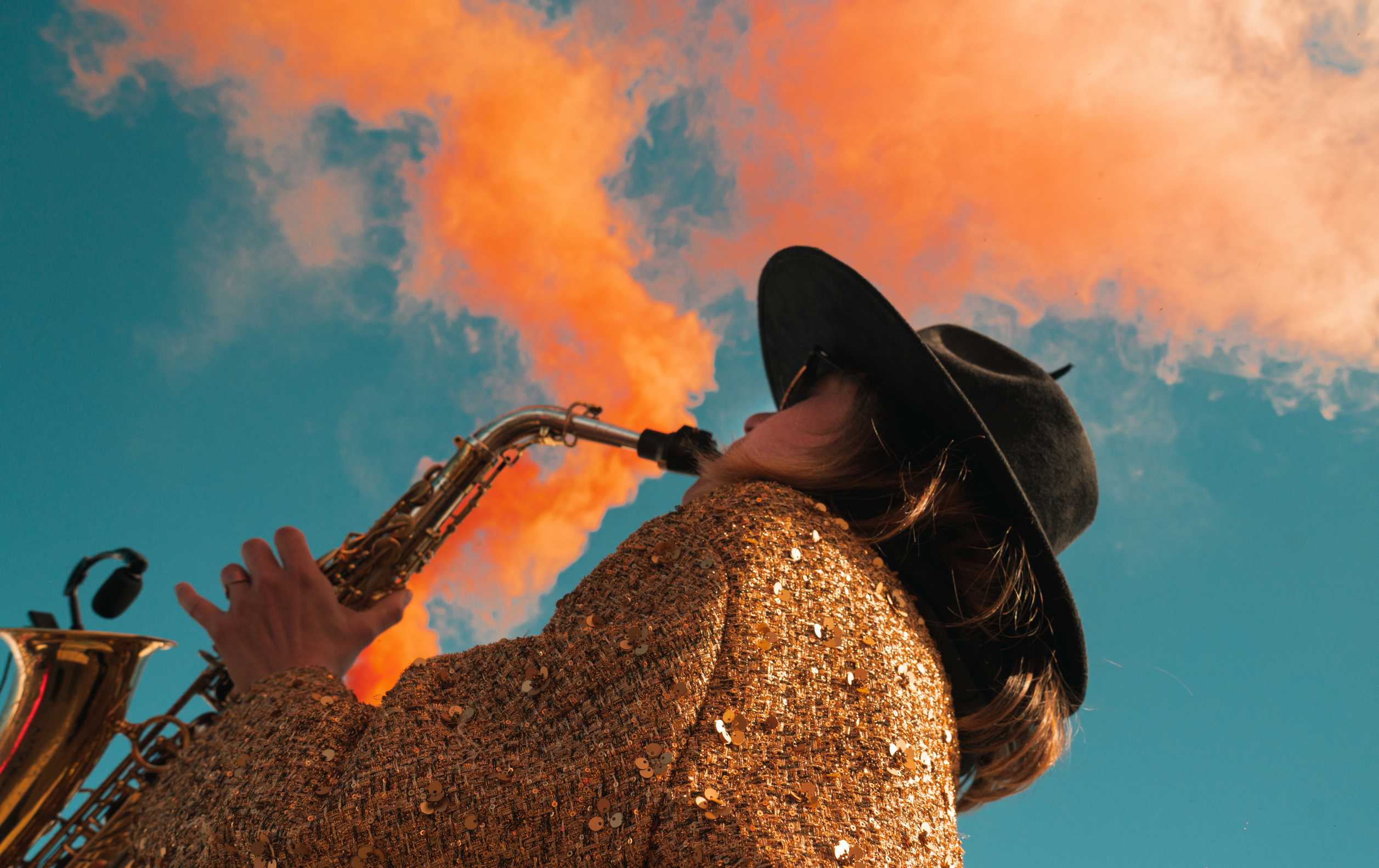 Person portant un chapeau noir et une veste à paillettes or joue du saxophone sous un ciel bleu avec des nuages orange.